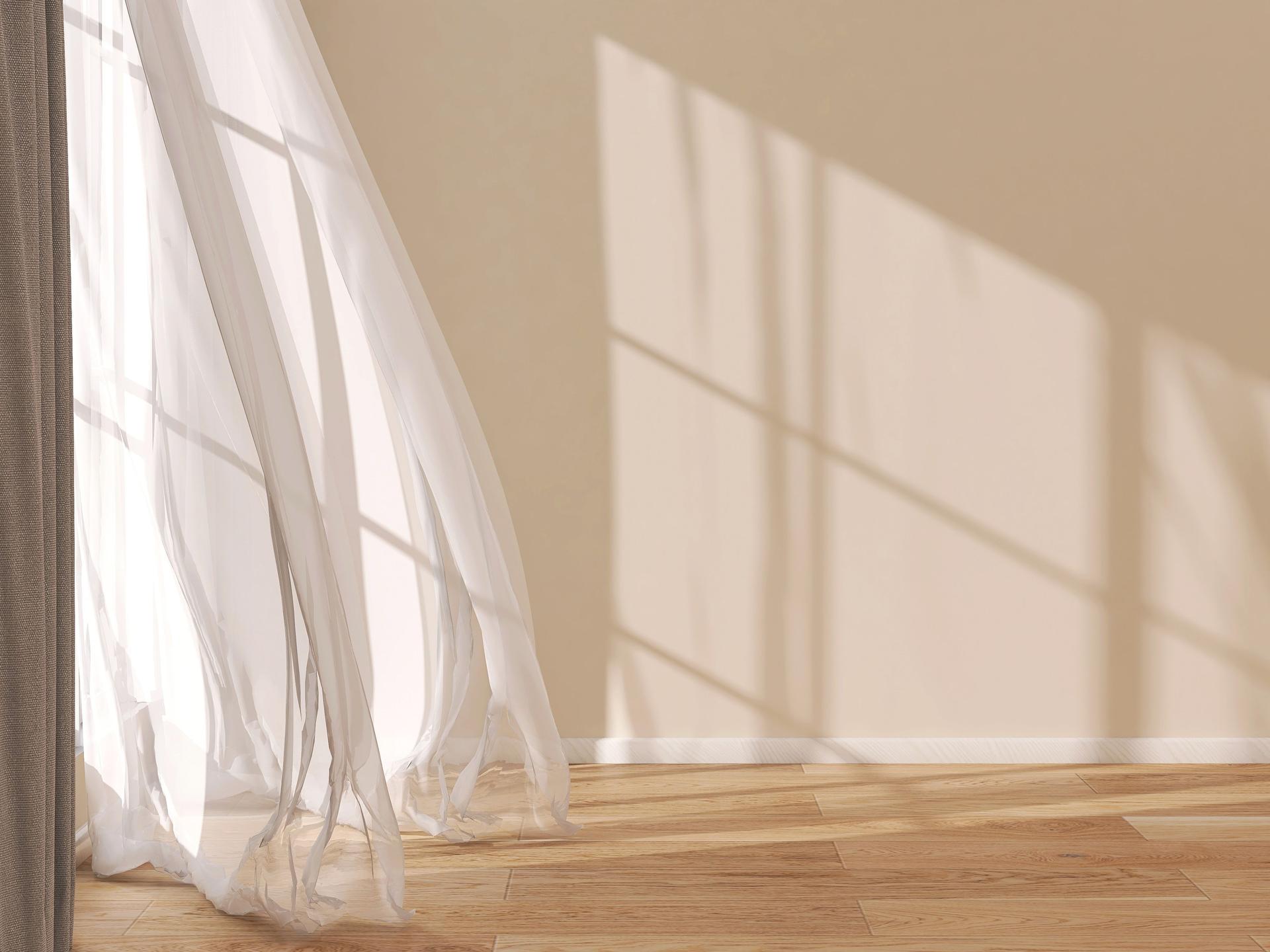 Front view of empty room with beige wall, white baseboard, blowing sheer curtain and wooden parquet floor in sunlight from window